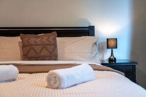 Ein ordentlich bezogenes Bett mit gestreifter Bettwäsche, einem braun gemusterten Kissen und einem aufgerollten weißen Handtuch. Ein Nachttisch mit einer Lampe steht neben dem Bett.