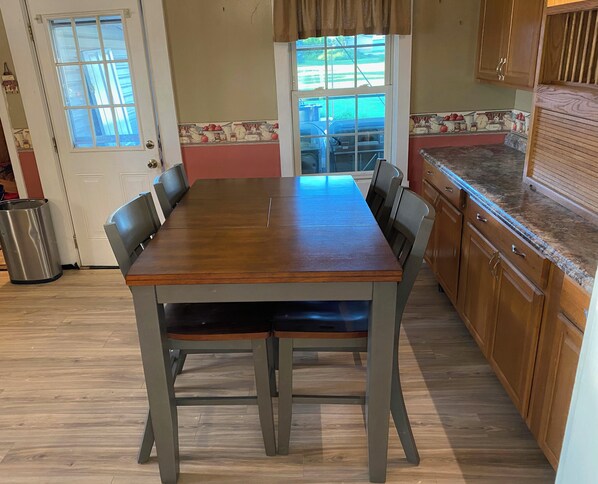 A dining area with a wooden table and chairs, a kitchen with wooden cabinets, and a window with a view of the outdoors.