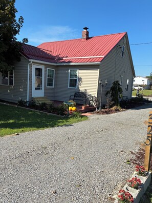A single-story house with a red roof, a front porch with a bench, and a gravel driveway.