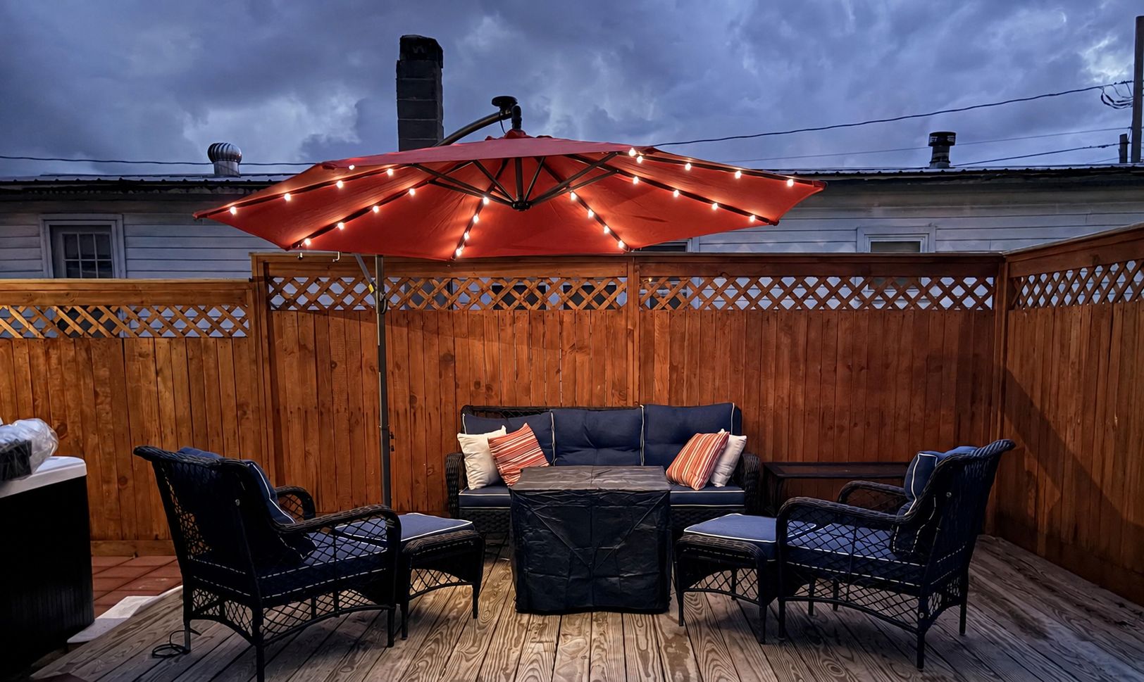 An outdoor patio with a wooden fence, a red umbrella with lights, and wicker furniture.