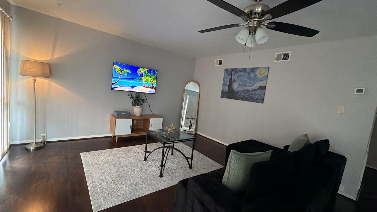 A living room with a flat-screen TV, a ceiling fan, a coffee table, a sofa, a mirror, and artwork on the walls.