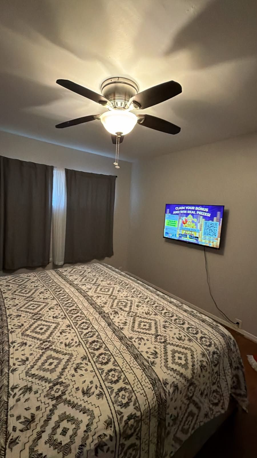 A bedroom with a bed, a ceiling fan, a television mounted on the wall, and a window with curtains.