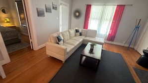 Ein Wohnzimmer mit einem beigefarbenen Ecksofa, einem Couchtisch und einem kleinen Beistelltisch. Eine Tür führt in einen anderen Raum, und ein Fenster mit roten Vorhängen ist vorhanden.