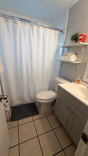 Un baño pequeño con cortina de ducha, inodoro, lavamanos y un estante con toallas.