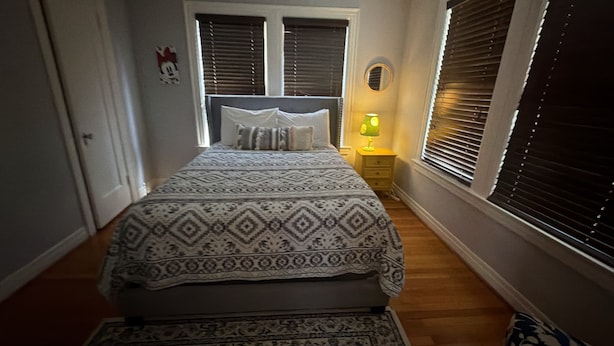 Ein Schlafzimmer mit einem Bett, einem Nachttisch mit Lampe, einem Fenster mit Jalousien und einer Tür.