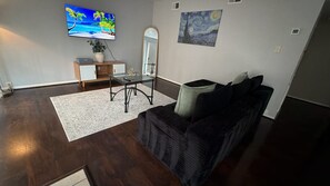 Una sala de estar con piso de madera oscura, una mesa de centro de vidrio, un sofá negro con cojines, un tapete blanco, una mesita de madera con una planta, un televisor de pantalla plana y un cuadro en la pared.