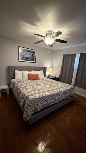 Habitación de hotel con cama, ventilador de techo, mesita de noche, lámpara, un cuadro enmarcado en la pared y una ventana con cortinas.