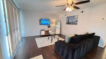 A living room with a dark-colored sofa, a small table with a lamp, a painting, and a ceiling fan.