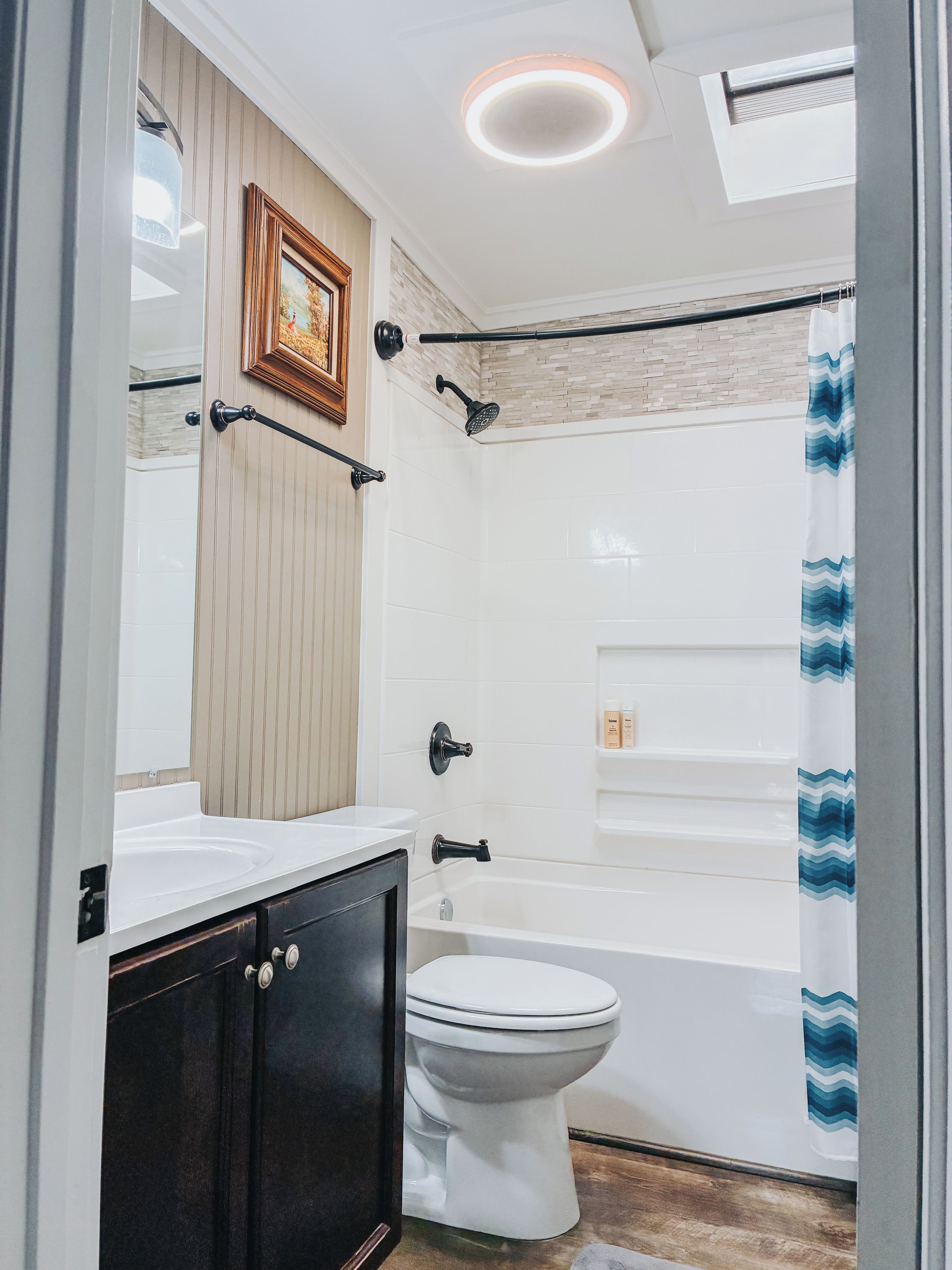 A bathroom with a white bathtub, a toilet, a vanity with dark cabinets, a framed picture on the wall, and a shower curtain with a blue pattern.
