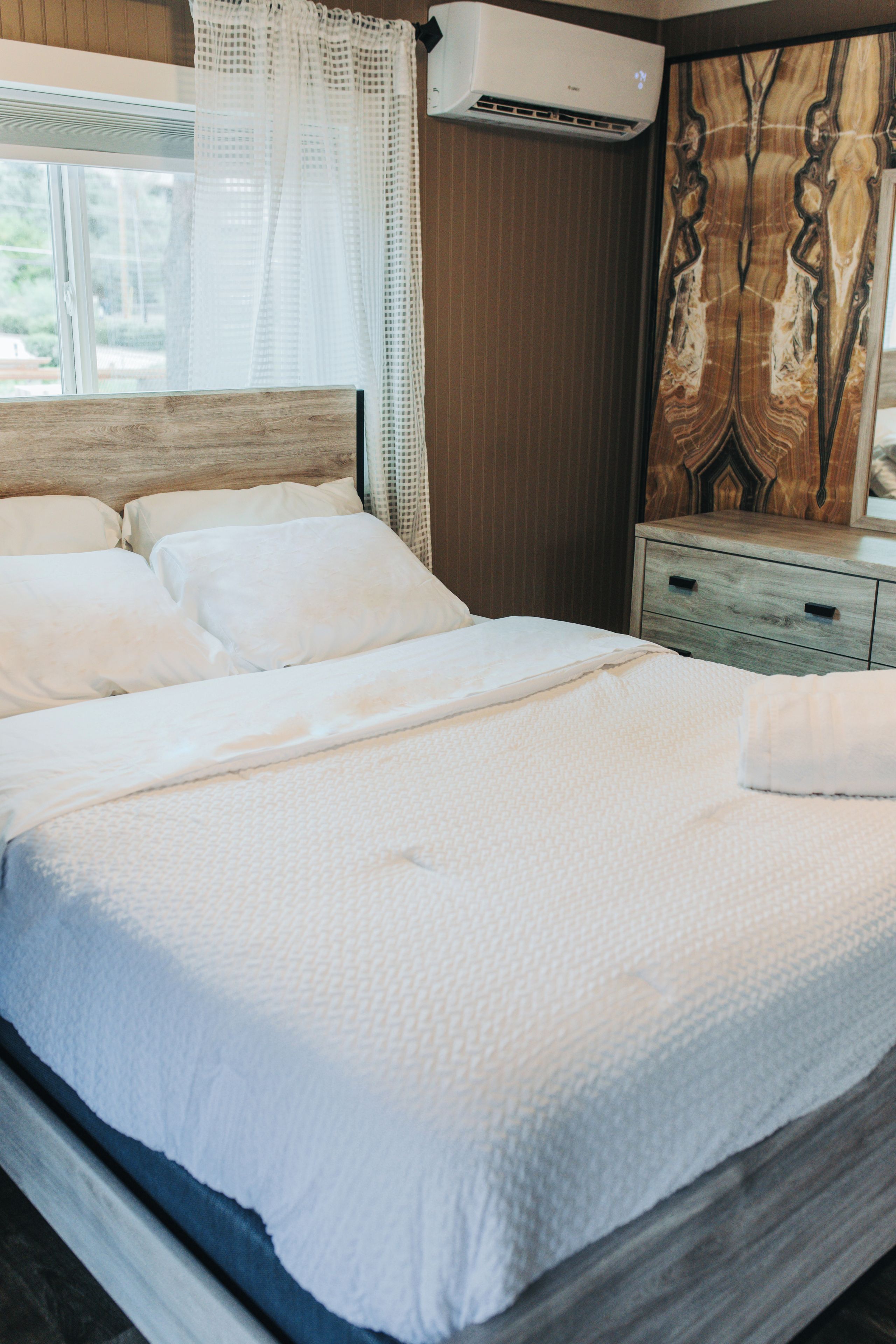 A neatly made bed with white linens, a wooden headboard, and a wall-mounted air conditioner.