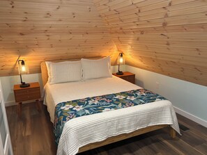 A bedroom with a wooden sloped ceiling, a bed with a floral bedspread, two bedside lamps, and a nightstand with a drawer. - Adagio 2: Luxury 2-bd 1-ba apt with stunning views close to Little River Canyon. (Gaylesville)