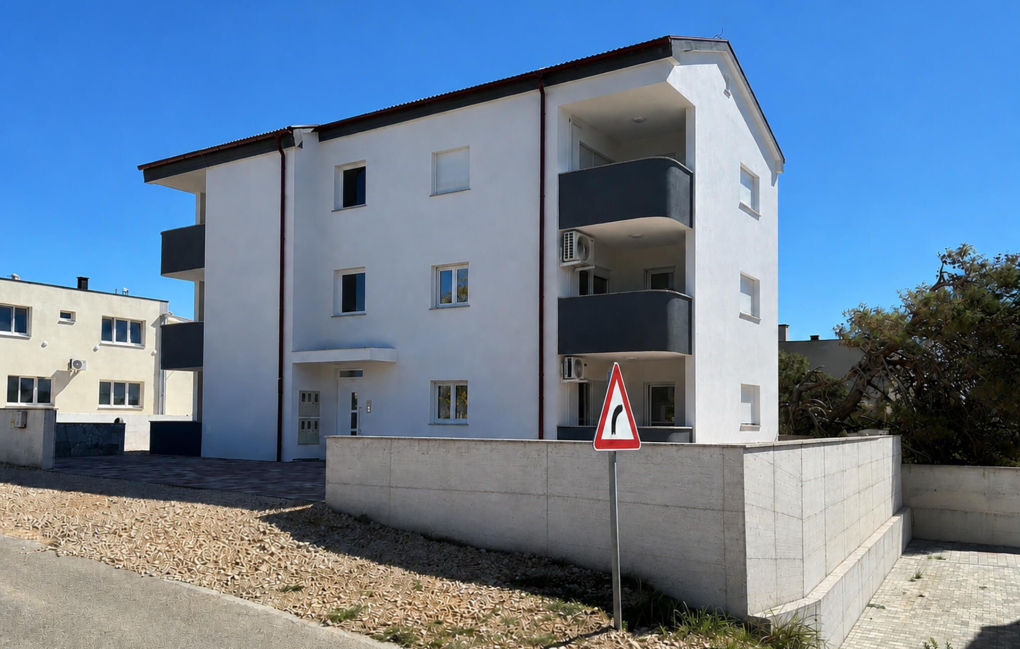 A modern multi-story residential building with balconies, a visible entrance, and a triangular warning sign.