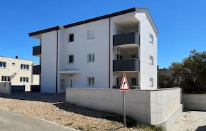 A modern multi-story residential building with balconies, a visible entrance, and a triangular warning sign. - 2 bedroom cozy apartment in Povljana (Povljana)