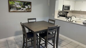 Un coin repas avec une table et des chaises, un ventilateur de plafond, une cuisine avec des meubles blancs et un tableau encadré au mur.