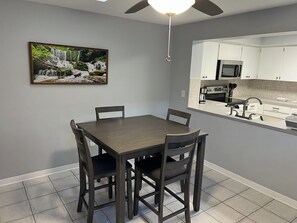 Un comedor con una mesa y sillas, un ventilador de techo, una cocina con armarios blancos y un cuadro en la pared.