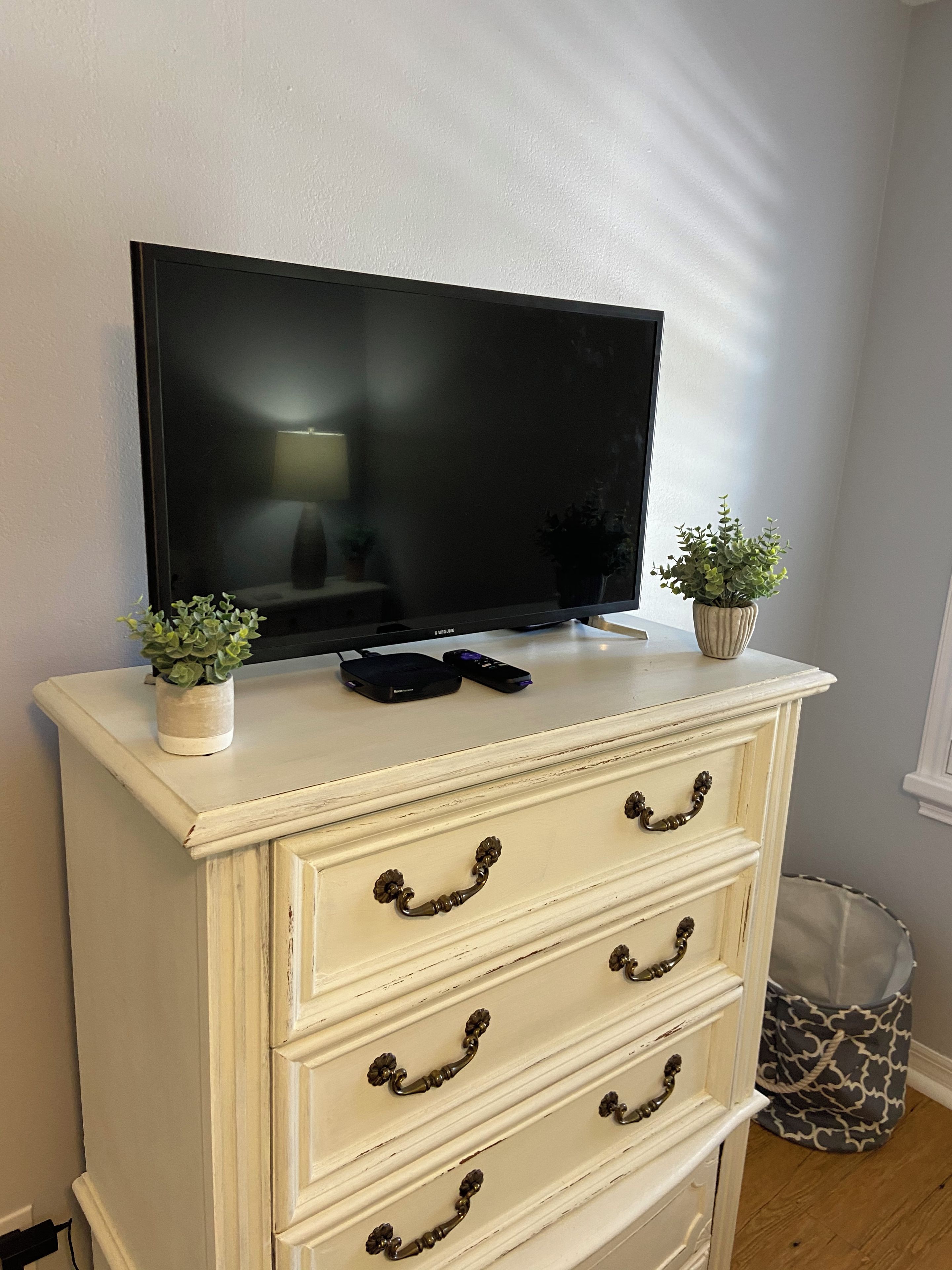 A white dresser with three drawers, a television on top, and two small potted plants.