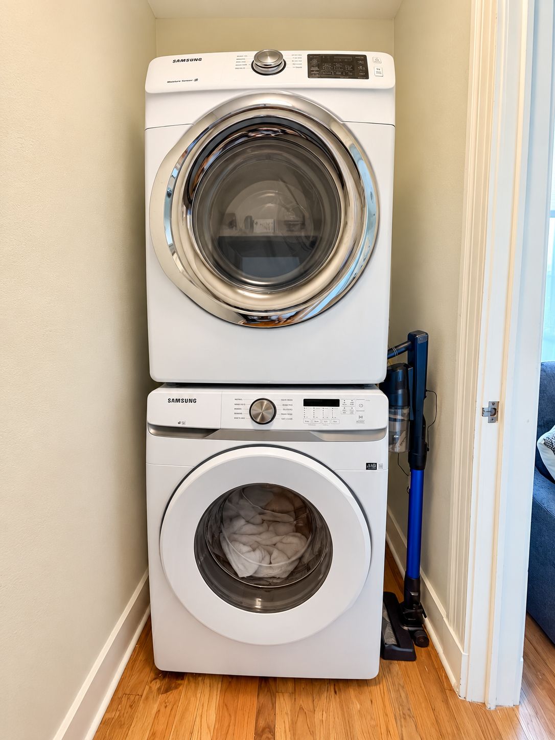 A Samsung front-loading washer and dryer stacked in a laundry room with a blue vacuum cleaner beside them.