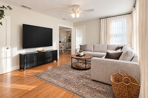 A modern living room with a flat-screen TV, a grey sofa, a coffee table, and a patterned rug. - LSU Gameday Cottage - Sleeps 12 - Private Gated Patio/Deck/Firepit (Baton Rouge)