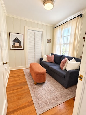 A room with a dark-colored sofa, a patterned rug, a wooden floor, a door with a light fixture above it, and a framed picture on the wall. - LSU Gameday Cottage - Sleeps 12 - Private Gated Patio/Deck/Firepit (Baton Rouge)