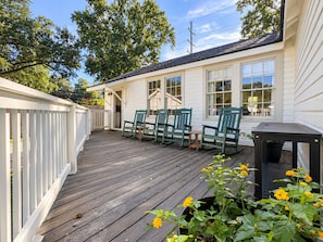 A wooden deck with outdoor seating, a white building with windows, and a tree in the background. - LSU Gameday Cottage - Sleeps 12 - Private Gated Patio/Deck/Firepit (Baton Rouge)
