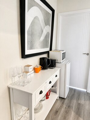 A white console table with a framed abstract painting, a coffee maker, a microwave, and a tea box.