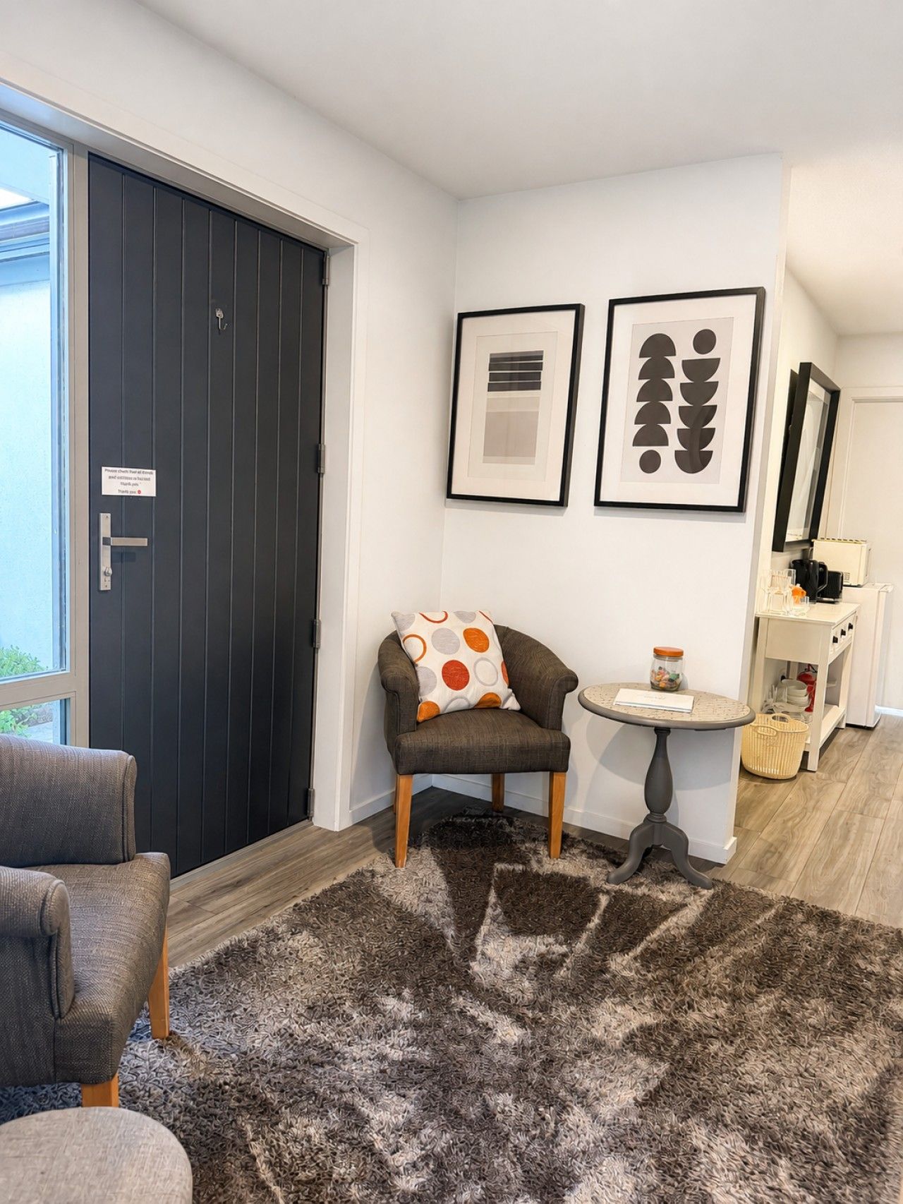 Een moderne kamer met een donkere deur, een grijze fauteuil, een klein rond tafeltje en twee ingelijste kunstwerken aan de muur.