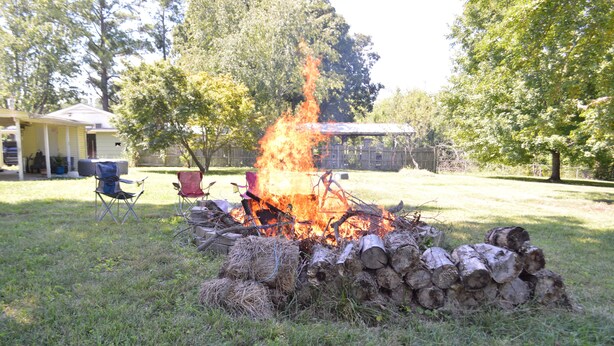 Un fuoco da campo con legna e rami, circondato da una zona erbosa con alberi e una casa sullo sfondo.