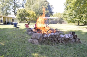A campfire with logs and branches, surrounded by a grassy area with trees and a house in the background.