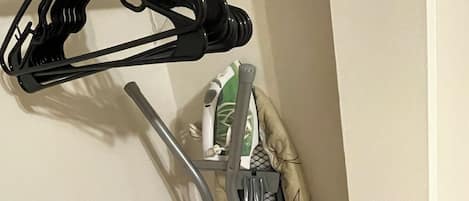 Esquina de una habitación con una viga de madera, un perchero negro y una tabla de planchar plegada apoyada en la pared.