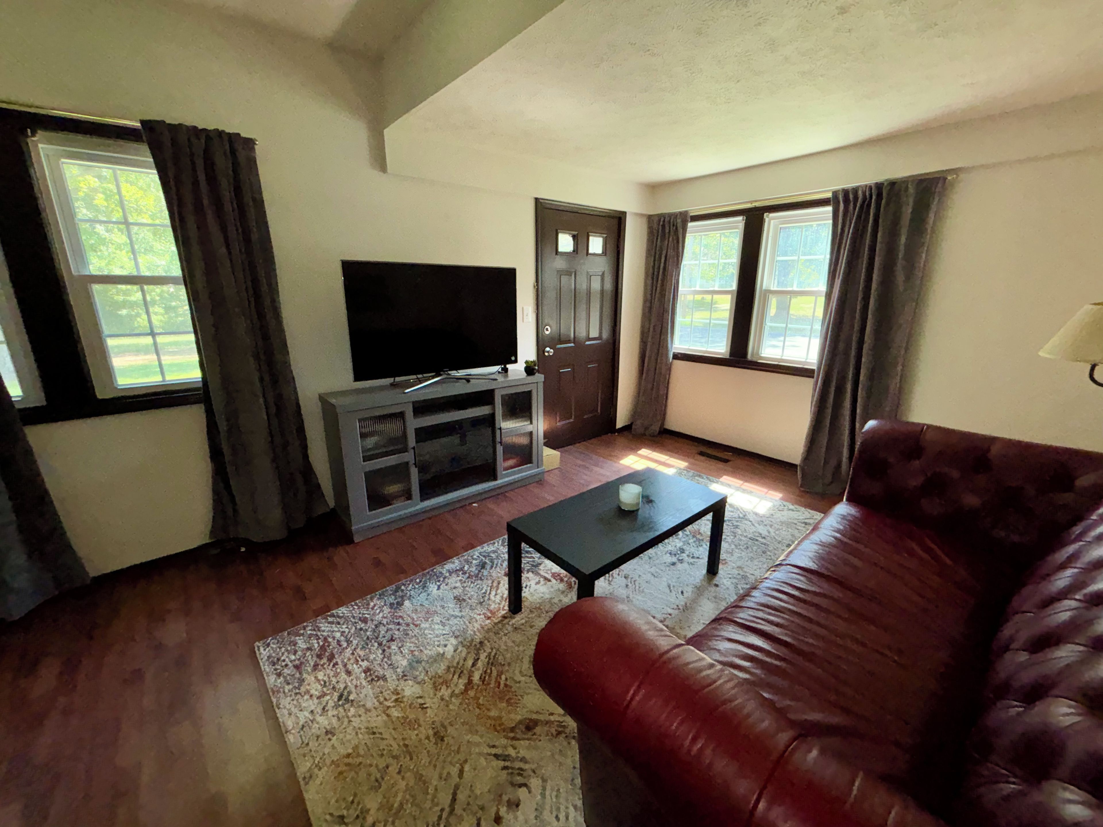 Una sala de estar con un sofá de cuero, un televisor sobre un mueble bajo y un ventanal con cortinas.