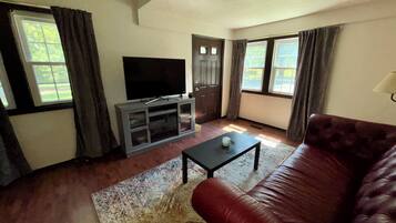 Una sala de estar con un sofá de cuero, un televisor sobre un mueble bajo y un ventanal con cortinas.