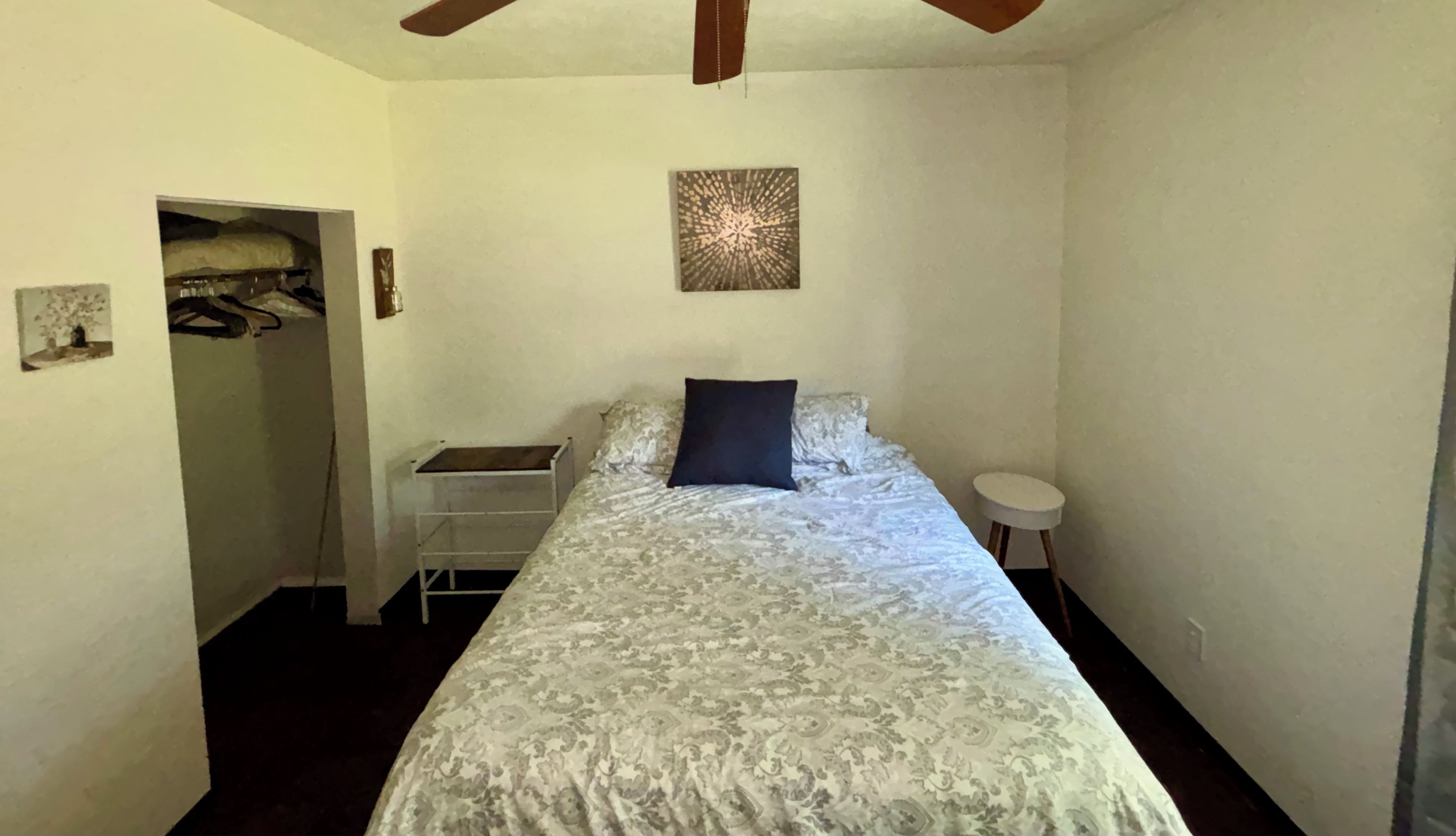 Une chambre à coucher avec un lit, une petite table, une chaise, une armoire et un ventilateur de plafond.