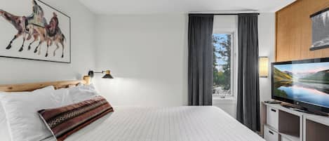 A hotel room with a bed, a mounted television, a painting of horses, a window with curtains, and a view of a mountain landscape.