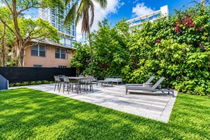 A patio with a table and chairs, surrounded by lush greenery and a tall building in the background. - 5 Bedroom | Home Near Wynwood | Pool |Free Parking (Miami)