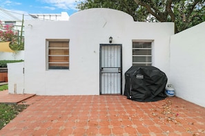 A white building with a black door, two windows, and a barbecue grill.