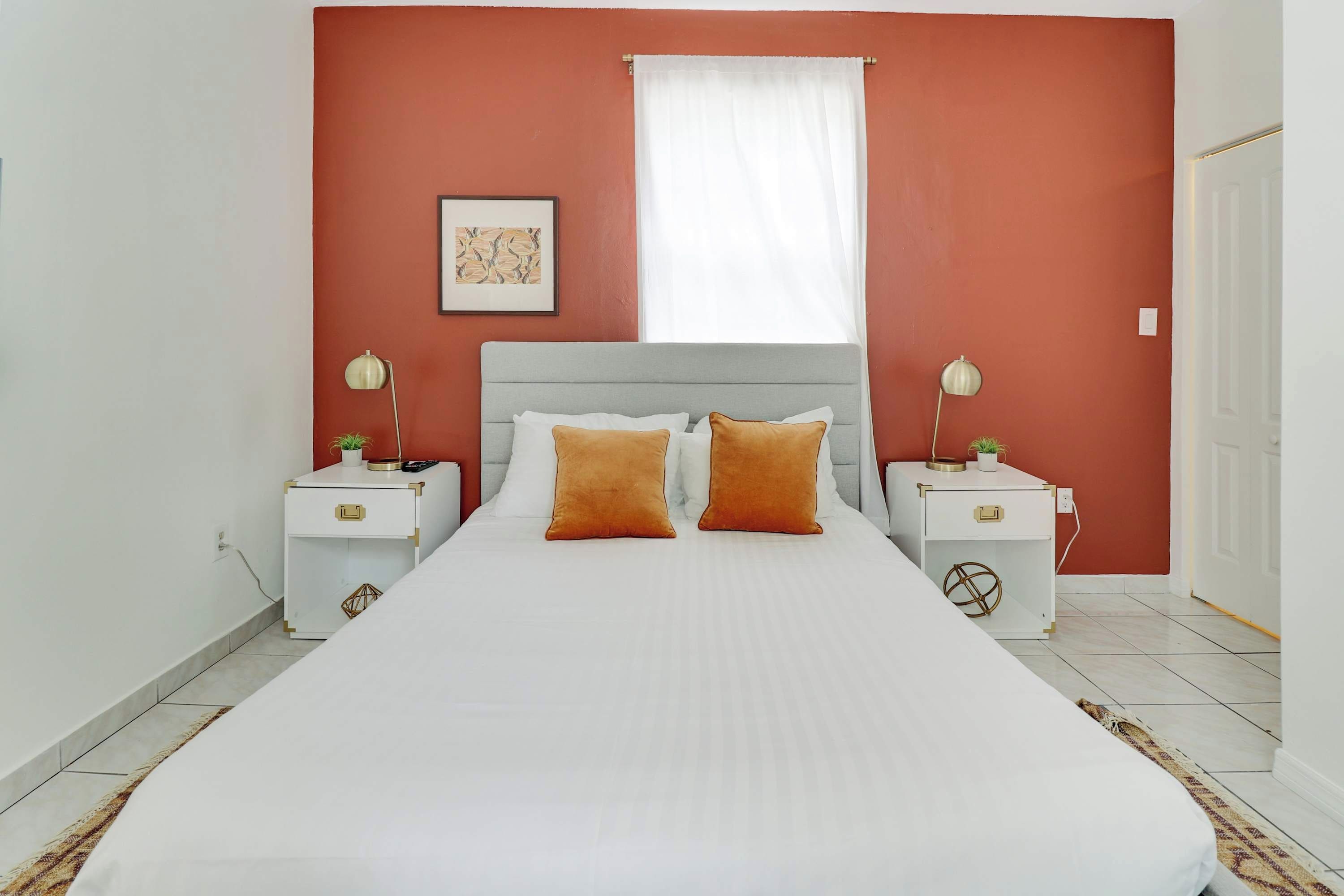 A bedroom with a large bed, two bedside tables, a window with white curtains, and a wall-mounted picture.