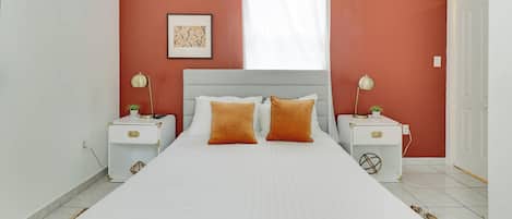 A bedroom with a large bed, two bedside tables, a window with white curtains, and a wall-mounted picture.