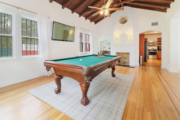 A spacious room with a pool table, wooden flooring, a ceiling fan, and a kitchen area visible through an open doorway.