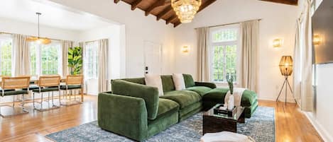 A spacious living room with a green sectional sofa, wooden flooring, and a high ceiling with exposed beams.