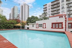 A swimming pool with a wooden deck, surrounded by a building with red window frames and a white facade.