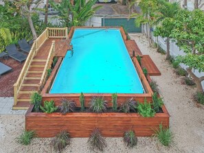 A rectangular swimming pool with a wooden deck and steps leading into it, surrounded by a garden with various plants.