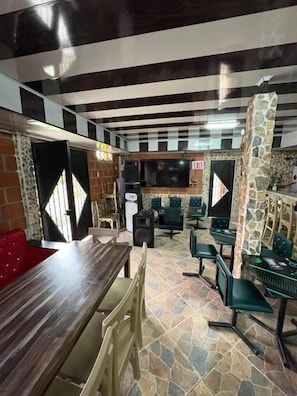 Um restaurante com uma longa mesa de madeira, várias cadeiras, uma tela de televisão e um pilar de pedra.