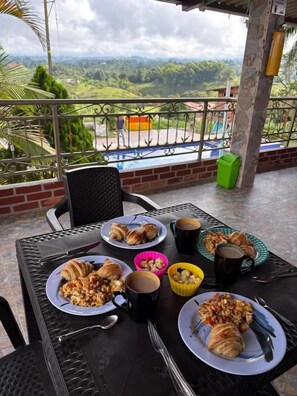 Une table à manger, dressée avec des assiettes remplies de nourriture, des tasses et des couverts, sur un balcon offrant une vue sur le paysage.