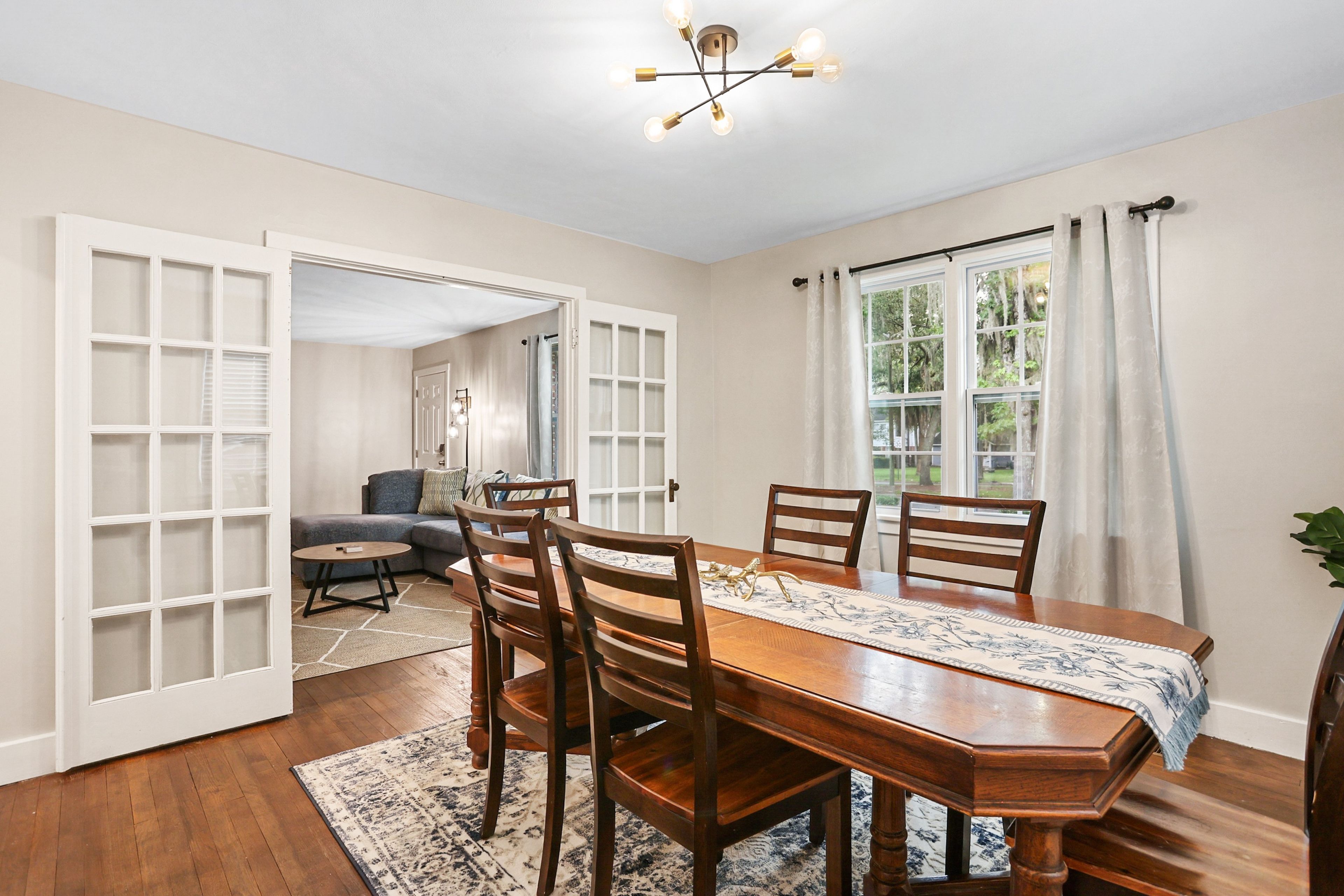 A dining room with a wooden table and chairs, a chandelier, and a view of the outdoors through a window.