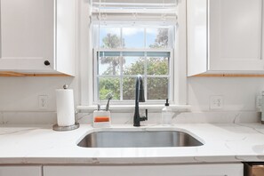 Eine Küche mit einer weißen Arbeitsplatte, einem Edelstahl-Spülbecken, einem Fenster mit Blick ins Grüne und einem Wasserhahn.