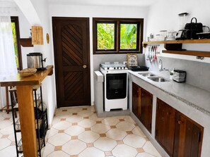 A compact kitchen with a stove, oven, and sink, wooden cabinets, and a window with a view of greenery. - Villaggio Arcobaleno (São Sebastião)