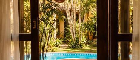 Zona de piscina interior con vista a un jardín tropical, un banco de madera y una mesa.