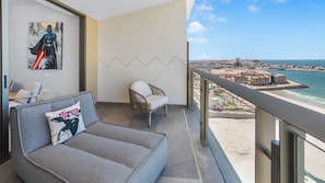 Een balkon met uitzicht op strand en stadsbeeld, een grijze bank met een kussen en een rieten stoel.