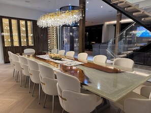 Una zona de menjador moderna amb una taula llarga, cadires blanques, un penjoll i una prestatgeria de vi.