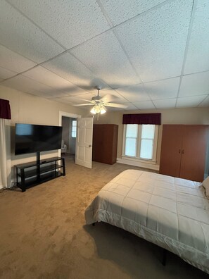 A bedroom with a bed, a TV on a stand, a ceiling fan, and a door leading to another room.